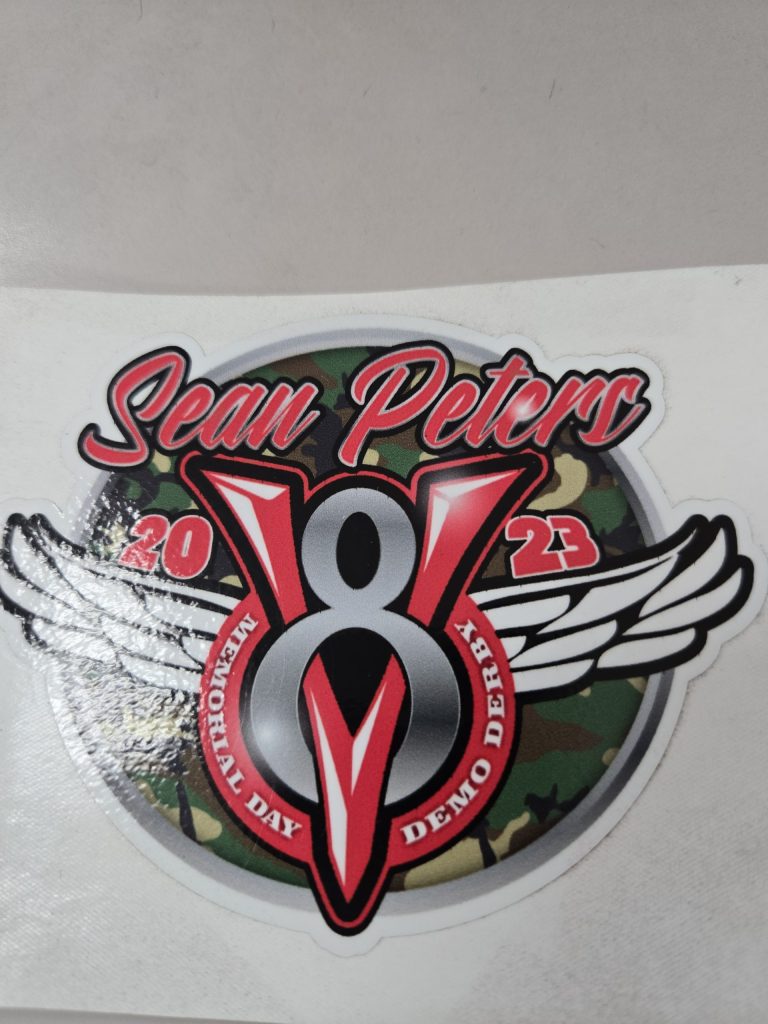 Sticker with "Sean Peters 2023 Memorial Day Demo Derby," featuring a V8 engine, wings, and camouflage background.