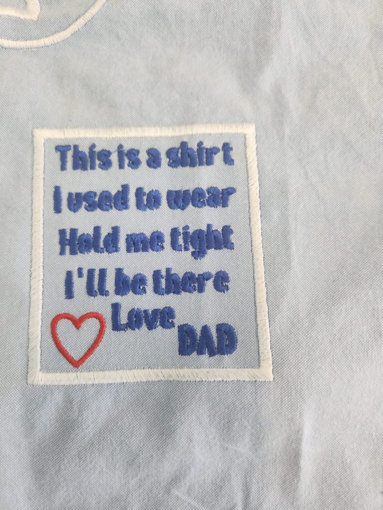 A patch on light fabric reads: "This is a shirt I used to wear. Hold me tight. I'll be there. Love, Dad" with a red heart.