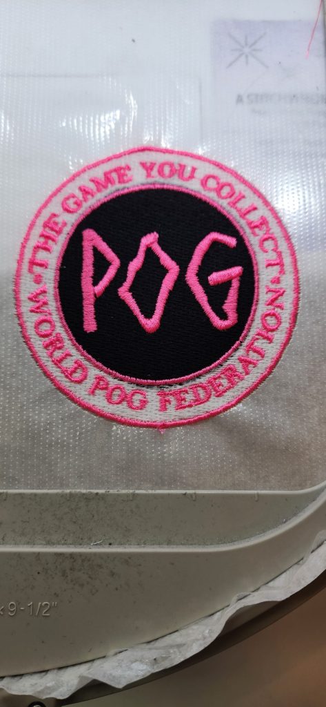 A circular patch reads "THE GAME YOU COLLECT, NOT PLAY, WORLD POG FEDERATION" with "POG" in pink letters at the center.