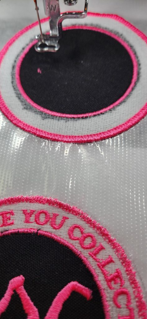 A sewing machine embroidering a circular black patch with pink and white thread on clear stabilizer.