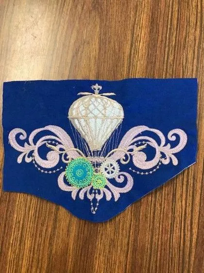 Blue fabric with an embroidered hot air balloon and decorative swirls, gears, and ornate designs on a wooden surface.