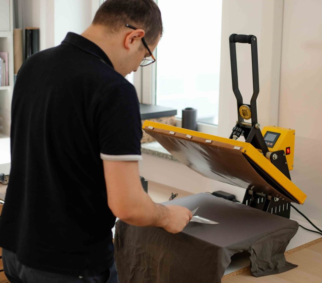A person uses a heat press machine to apply a design onto a dark t-shirt in a well-lit workspace.