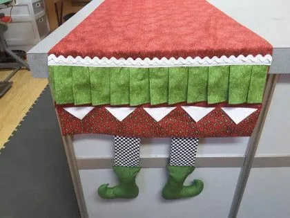 A table runner with a red, green, and white design, featuring elf legs and shoes hanging off the edge.