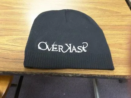 A black knit beanie with the word "Overkast?" embroidered in white on the front, lying on a wooden table.