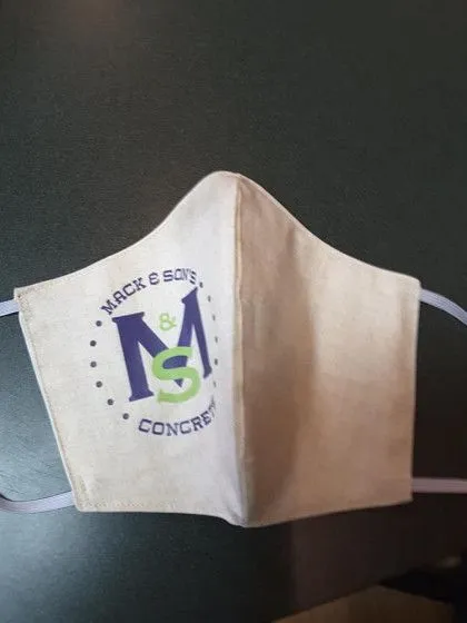 Beige cloth face mask with "Mack & Sons Concrete" logo in blue and green, lying on a dark surface.