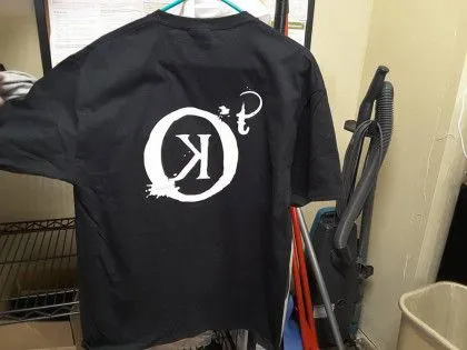 Black t-shirt hanging on a rack with a large white reversed "OK" symbol printed on the back.