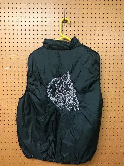 A black puffy vest with an embroidered wolf head design hangs on a yellow hanger against a pegboard wall.