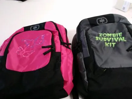 Two backpacks side by side: one pink with butterfly designs, one gray with "Zombie Survival Kit" text on the front pocket.