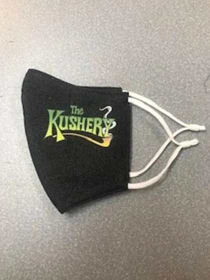 Black face mask with "The Kushery" logo in green and yellow, lying on a gray surface with white ear loops visible.