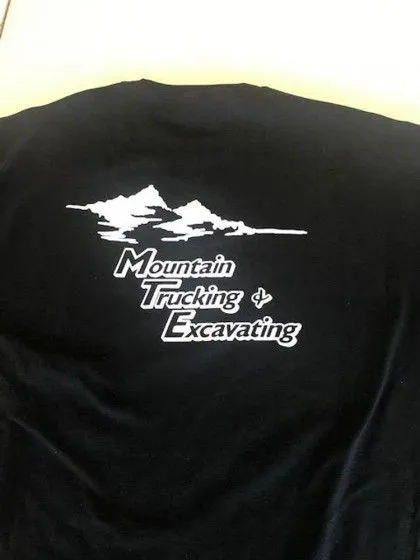 Black t-shirt with white graphic of mountains and text reading "Mountain Trucking & Excavating.