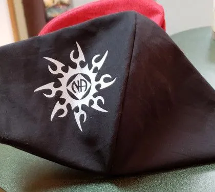 A black face mask with a white tribal sun and “NA” logo printed in the center lies on a green surface.