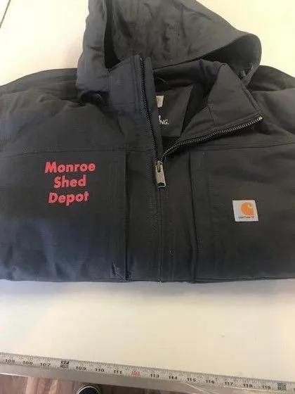 A dark hooded jacket with “Monroe Shed Depot” embroidered in red and a Carhartt logo patch on the pocket.