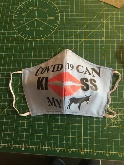 A cloth face mask with text "COVID 19 CAN KISS MY" featuring lips and a donkey graphic, on a green cutting mat.