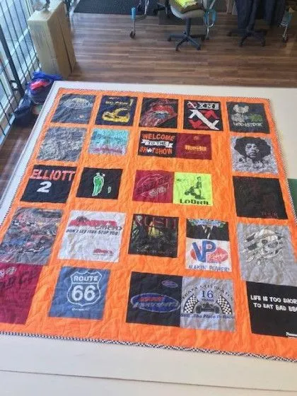 A quilt made from various colorful t-shirts laid out on a wooden floor near a window.