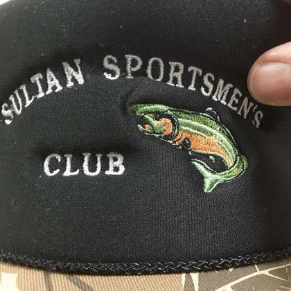 Black cap with "Sultan Sportsmen's Club" text and an embroidered fish, held by a person’s hand.