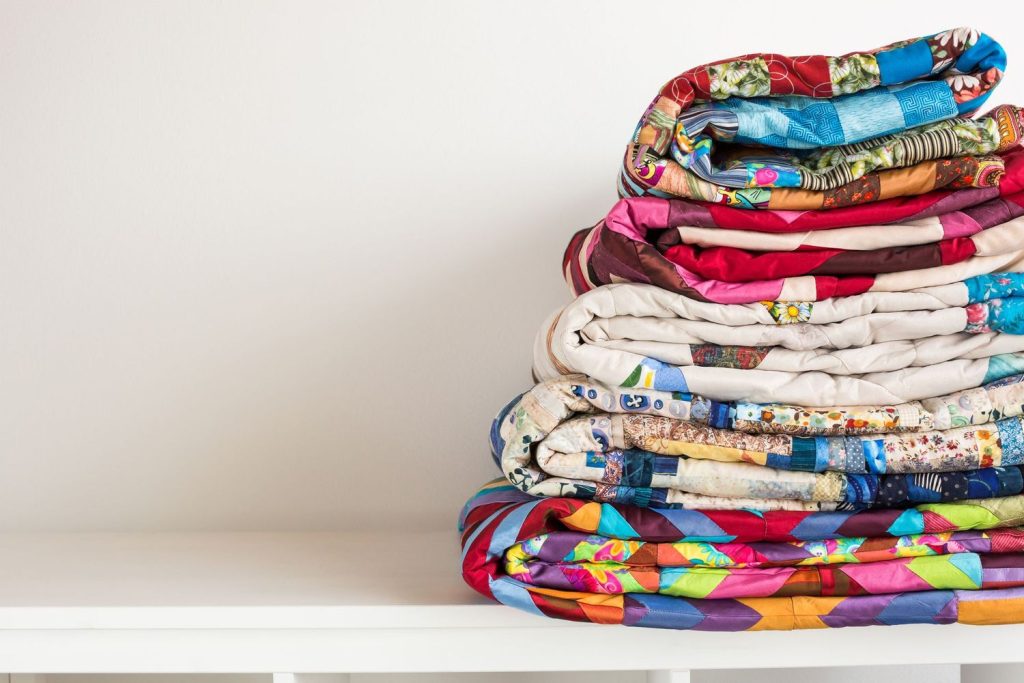 A stack of colorful folded quilts sits on a white surface against a plain white wall.
