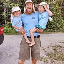 An adult holding two children, all wearing sun hats and blue shirts, standing on a dirt road by a wooded area.