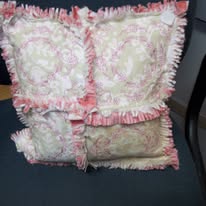 A square pillow with a pink and white floral pattern, divided into four sections with ruffled edges, on a dark chair.