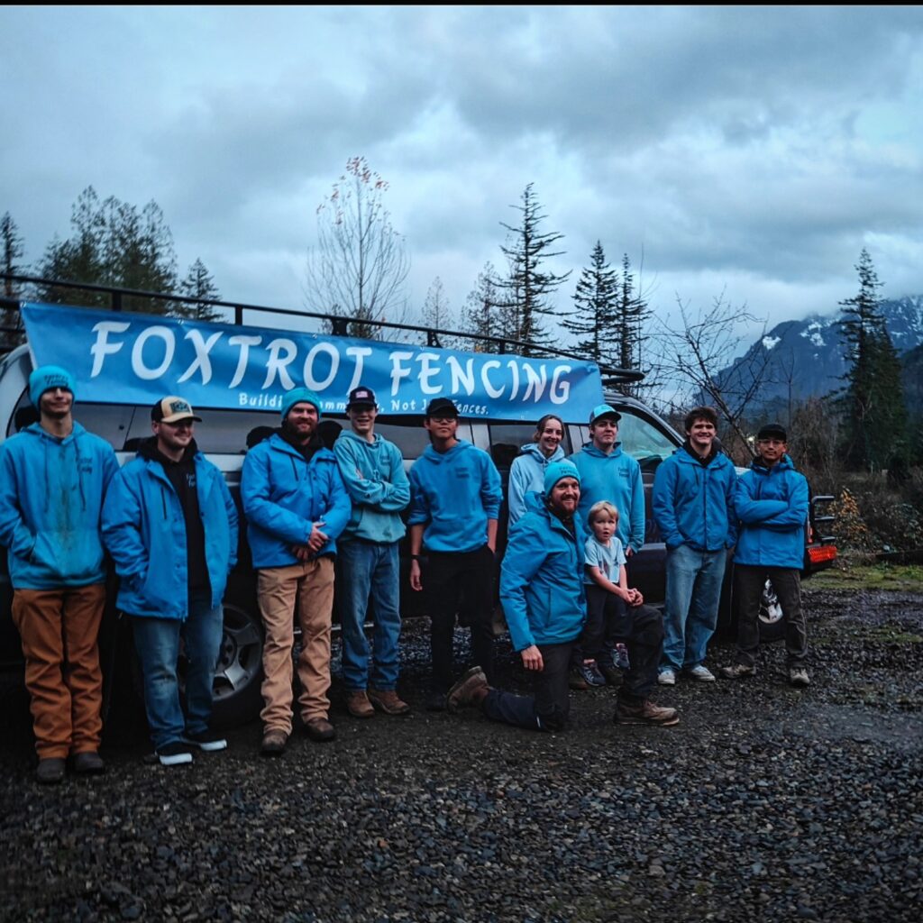 A group of people in blue jackets stand in front of a "Foxtrot Fencing" banner outdoors on a cloudy day.