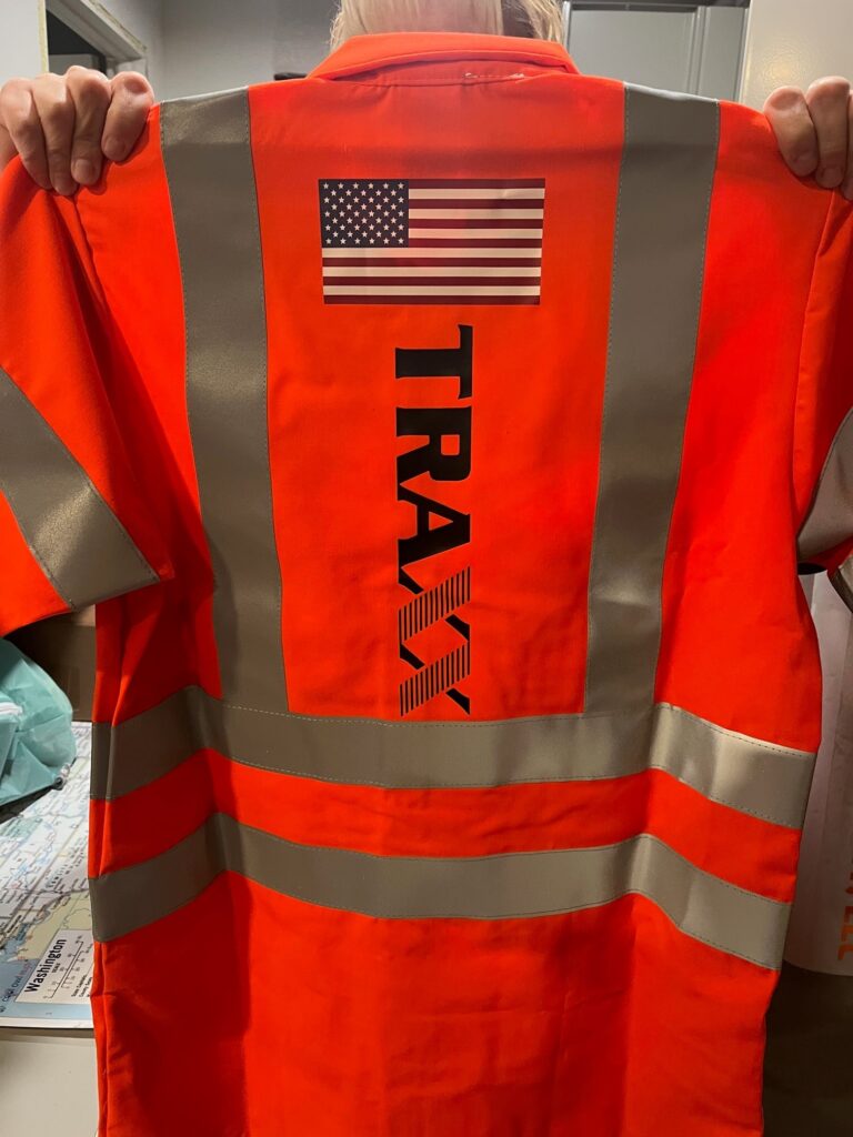 A person holds up an orange safety vest with reflective stripes, an American flag, and "TRAX" printed on the back.