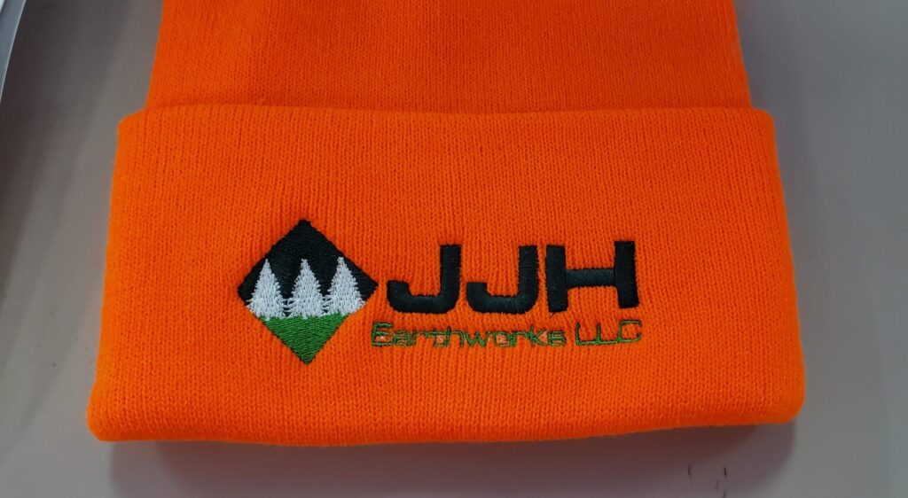 Orange knit hat with JJH Earthworks LLC logo embroidered in black, green, and white on the cuff.