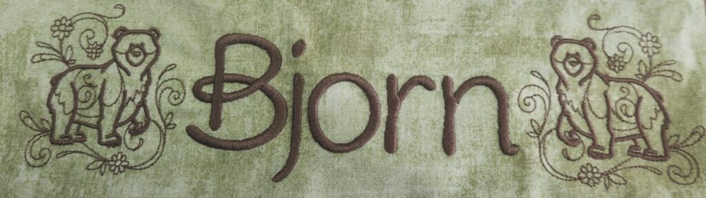 Embroidery of the name 'Bjorn' in brown thread on green fabric with bear and floral designs on each side.
